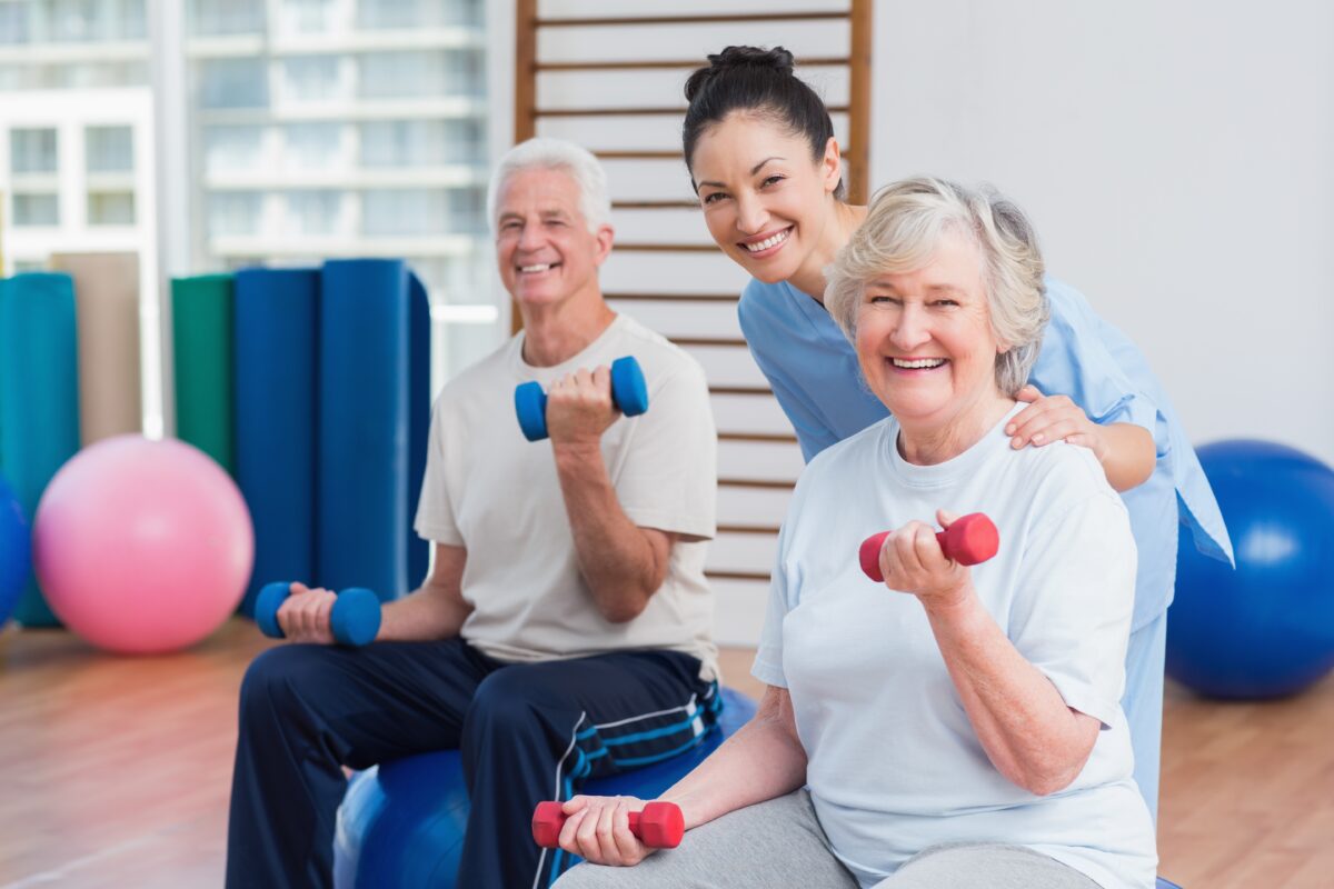 two older adults working out with barbells and a person supporting them and smiling in the background.